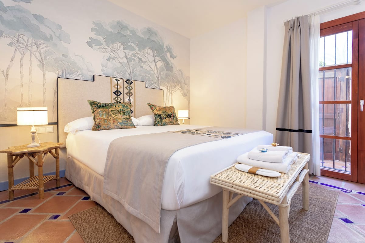 Una habitación de hotel con una cama grande, dos mesitas de noche con lámparas, una pequeña mesa con toallas y un mural en la pared.