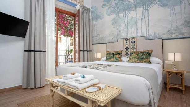 Una habitación de hotel con una cama, una mesita con toallas, un televisor montado en la pared y una ventana con cortinas.