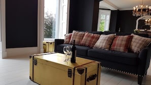 En stue med en mørkeblå sofa, ternede puder, et gulvtæppe-bord med vinglas og en flaske, samt en lysekrone.