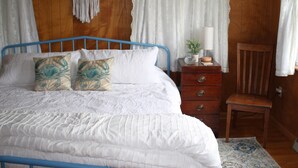 A bedroom with a blue metal bed frame, white bedding, two decorative pillows, a wooden nightstand with a lamp, and a wooden chair.