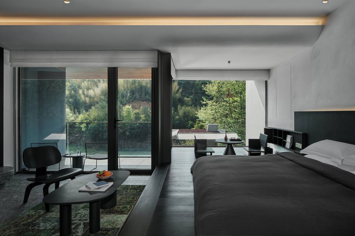 A modern bedroom with a large bed, a desk, and a chair. There is a balcony with a view of trees and a building.