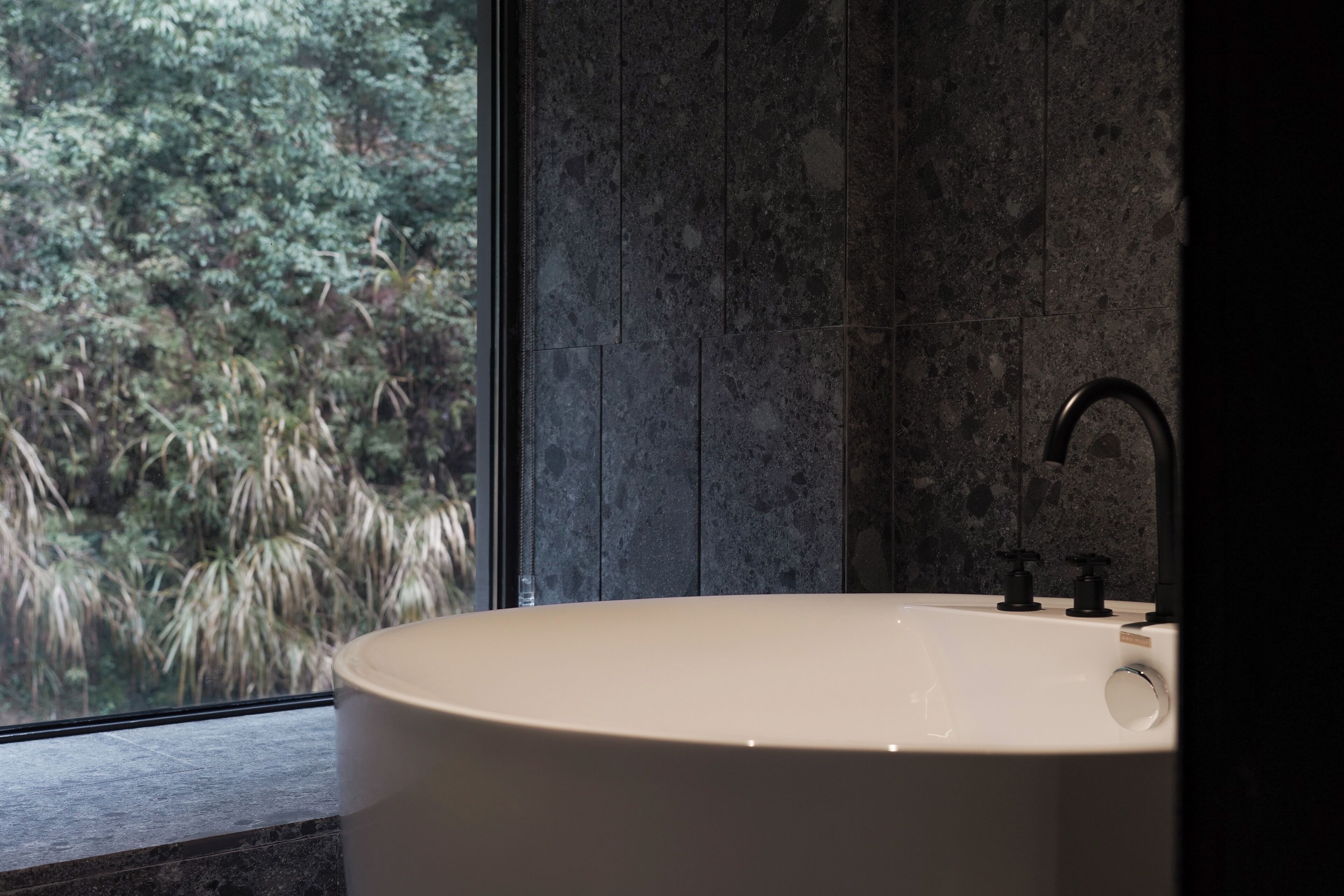 Un baño moderno con una bañera independiente, un ventanal con vistas a vegetación y una pared de azulejos oscuros.