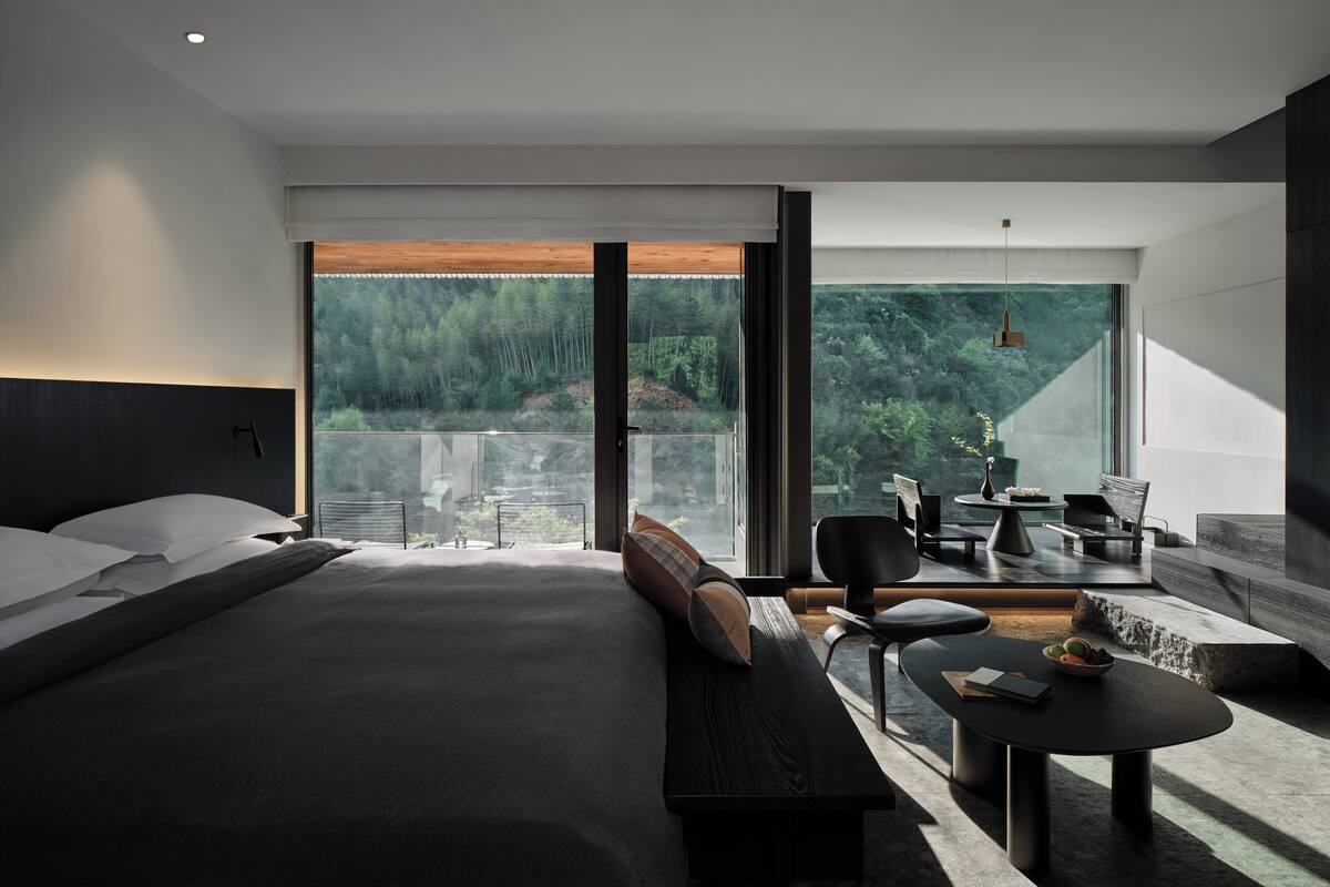 A modern hotel room with a large bed, a seating area with a table, and a balcony with a view of the forest.