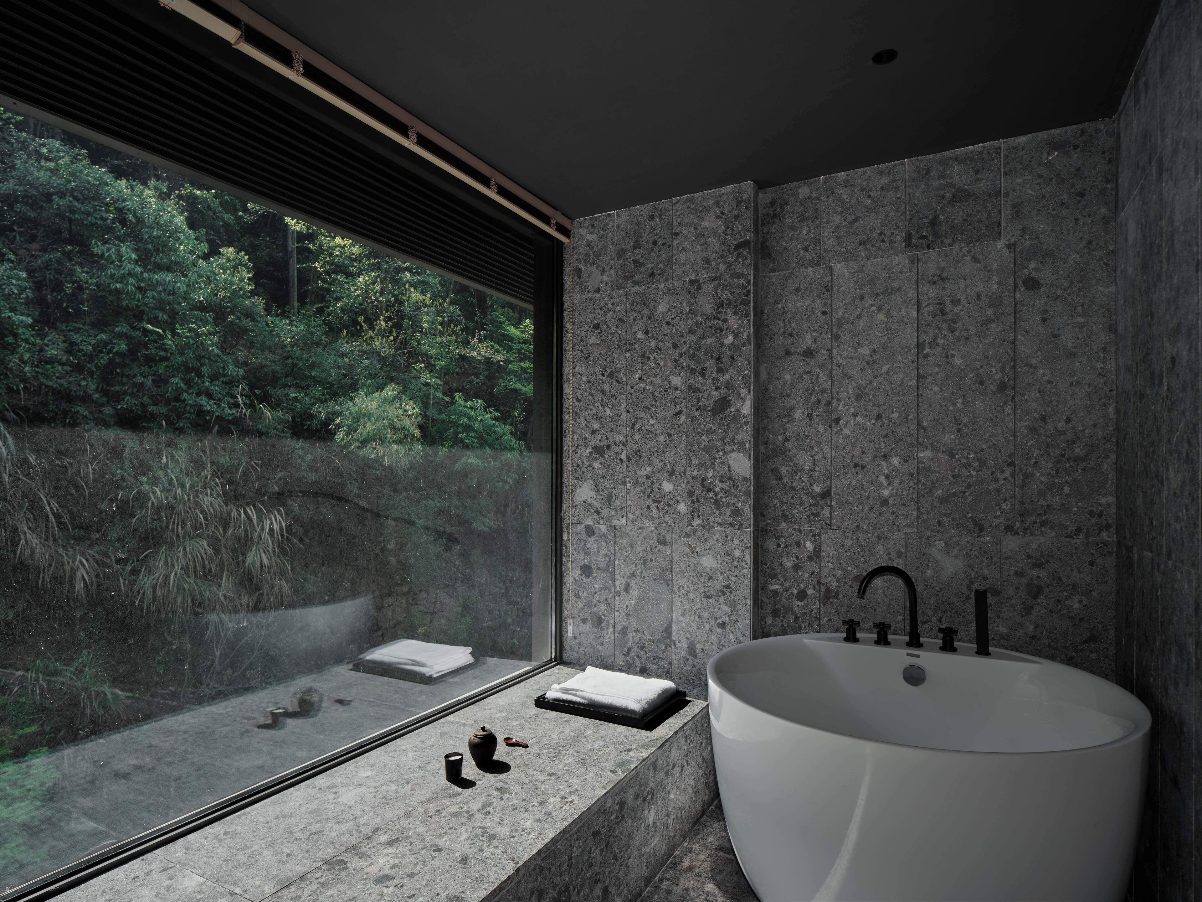 Un baño moderno con una bañera independiente grande, una ventana con vistas a follaje y una pared con textura de piedra.