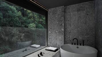 Un baño moderno con una bañera independiente grande, una ventana con vistas a follaje y una pared con textura de piedra.