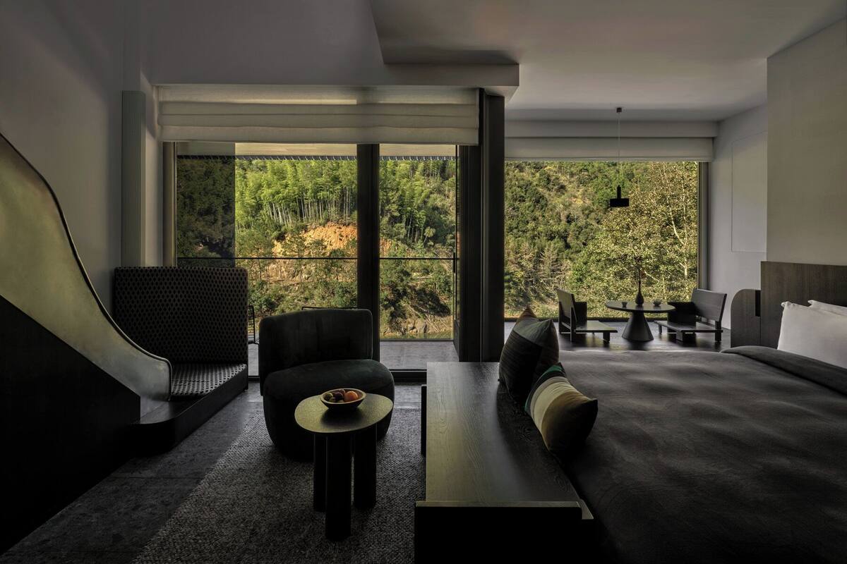 A modern hotel room with a large window offering a view of a forested area. There is a sofa, a small round table with a bowl of fruit, and a bed with a headboard.
