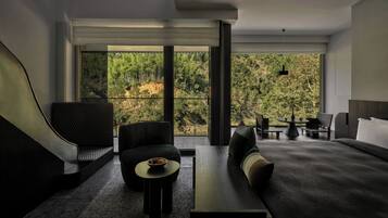 A modern hotel room with a large window offering a view of a forested area. There is a sofa, a small round table with a bowl of fruit, and a bed with a headboard.
