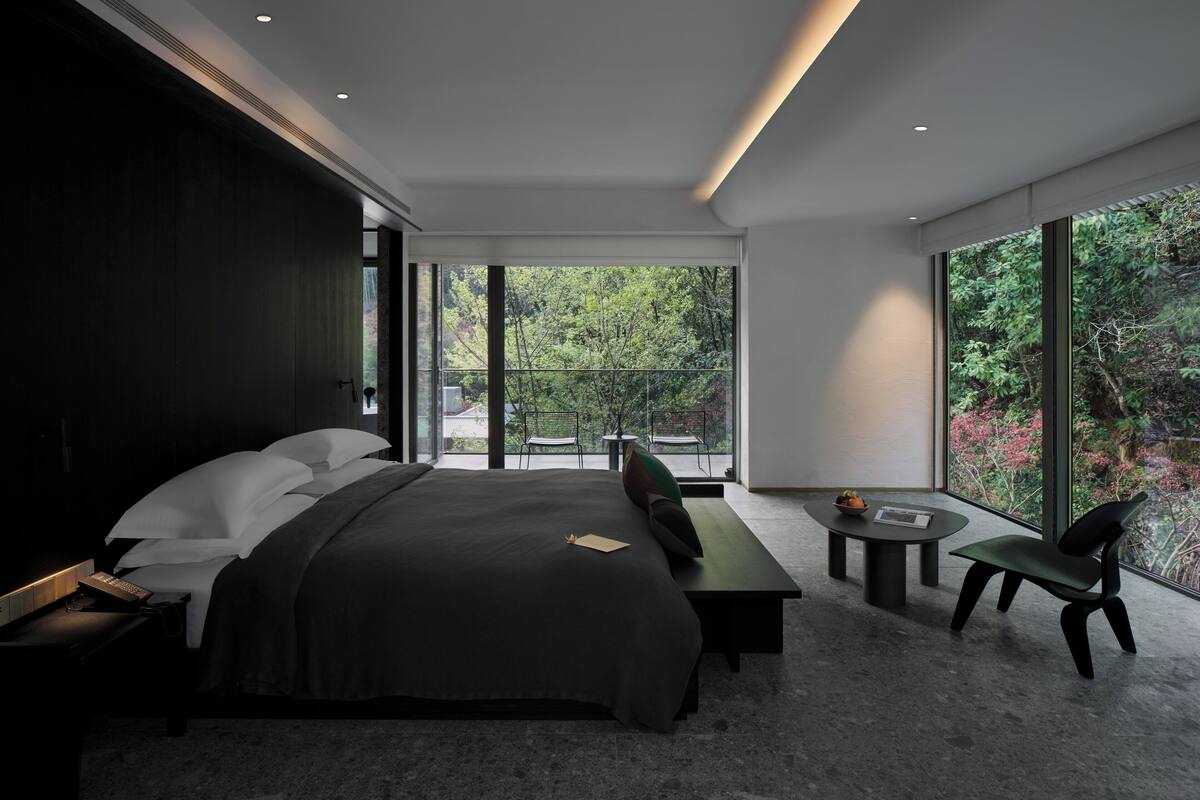 A modern bedroom with a large bed, a desk, and a chair, featuring a view of greenery through large windows.