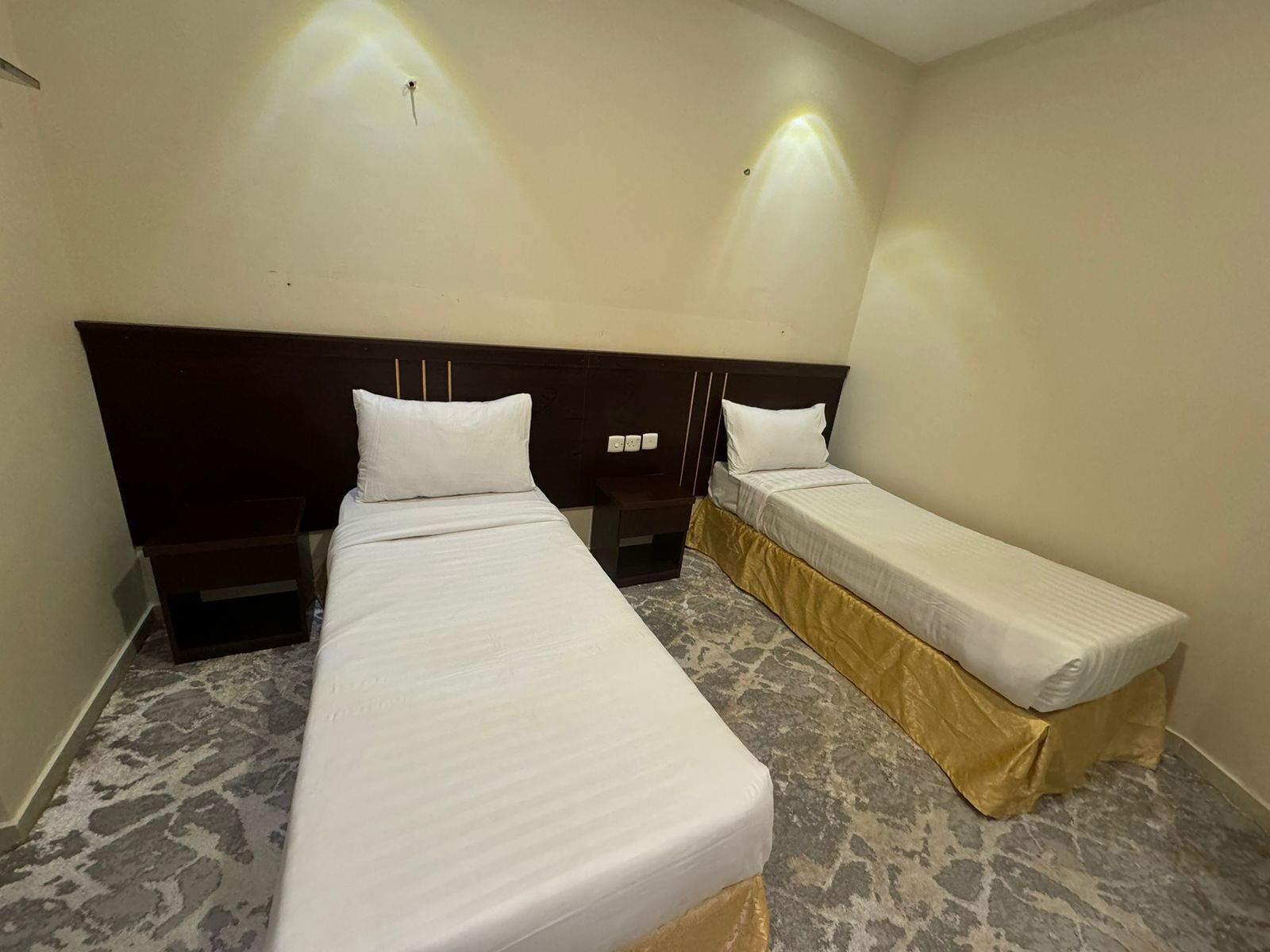 Una camera d'albergo con due letti singoli, una testata del letto scura, un pavimento effetto marmo e un lampadario a parete.