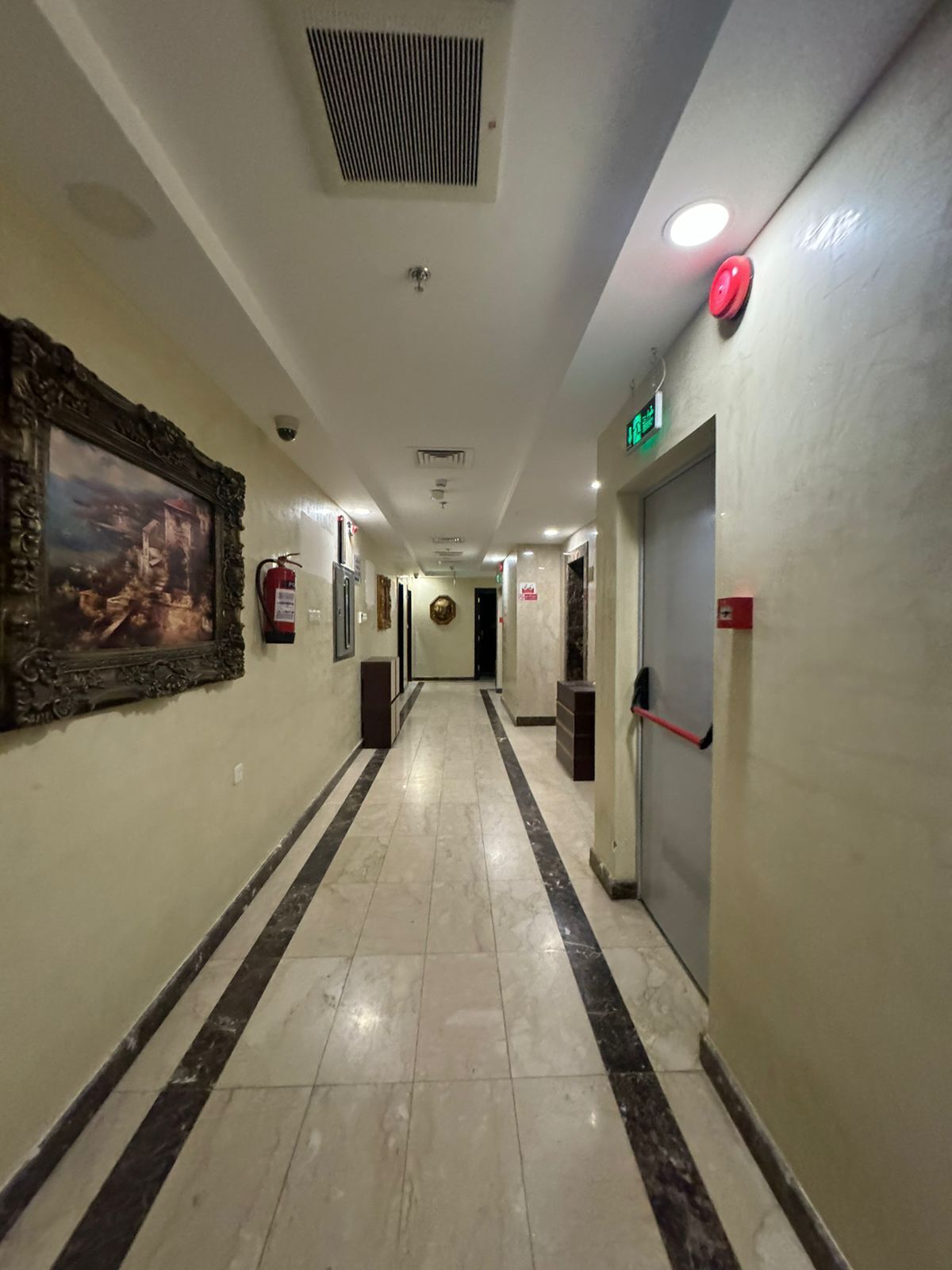 Ein Hotelkorridor mit Marmorboden, gerahmten Kunstwerken, einem Feuerlöscher und einem Notausgangsschild.