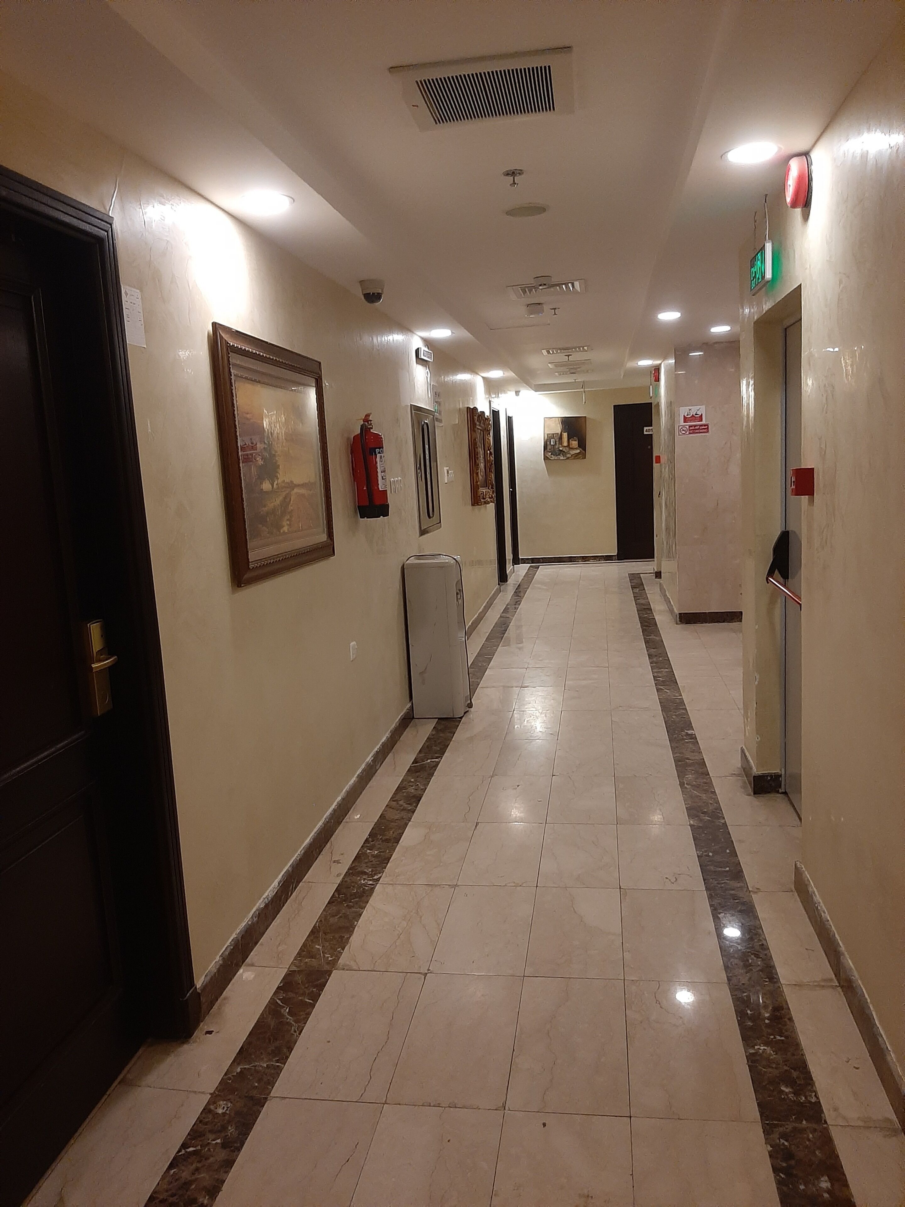A hotel corridor with a tiled floor, framed artwork, a fire extinguisher, and a digital clock.