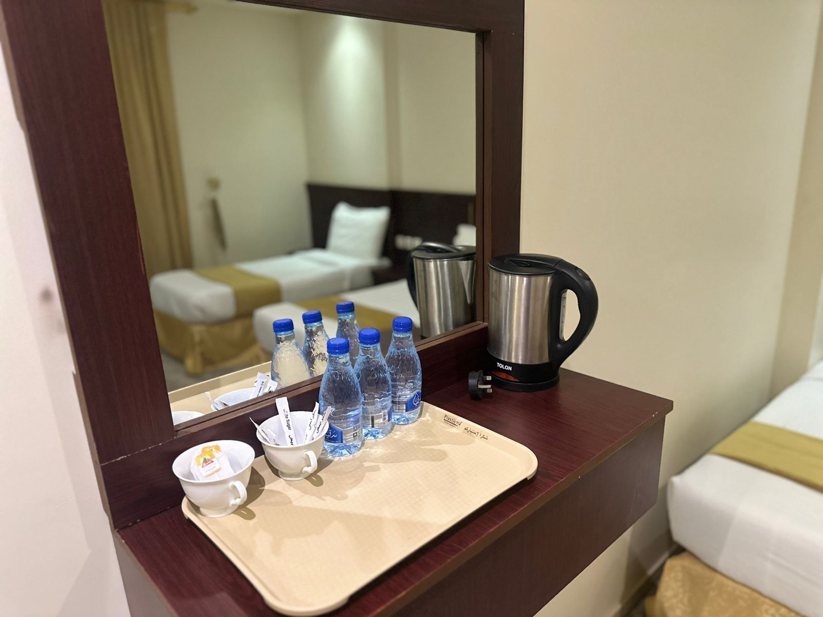 A hotel room with a wooden desk, a mirror, a bed, a coffee maker, and several water bottles.