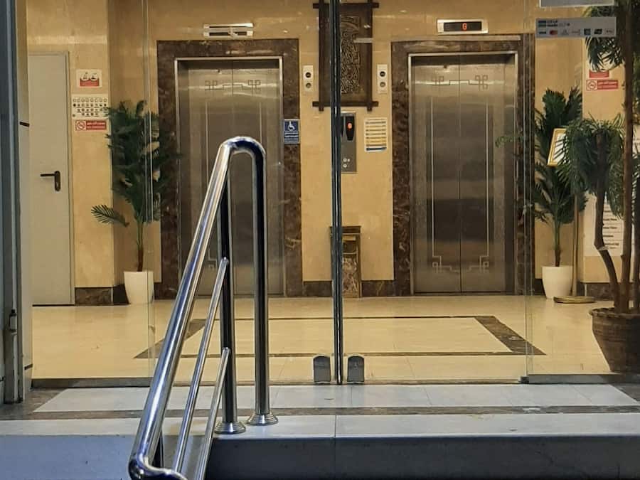 Entrada de hotel con puerta de cristal, dos ascensores, una araña y una escalera con barandillas de metal.
