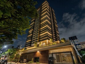 Un edificio de gran altura con múltiples balcones, entrada protegida y zona de estacionamiento.