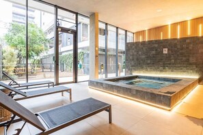Un área de piscina interior moderna con paredes de vidrio, un jacuzzi y sillones de descanso.