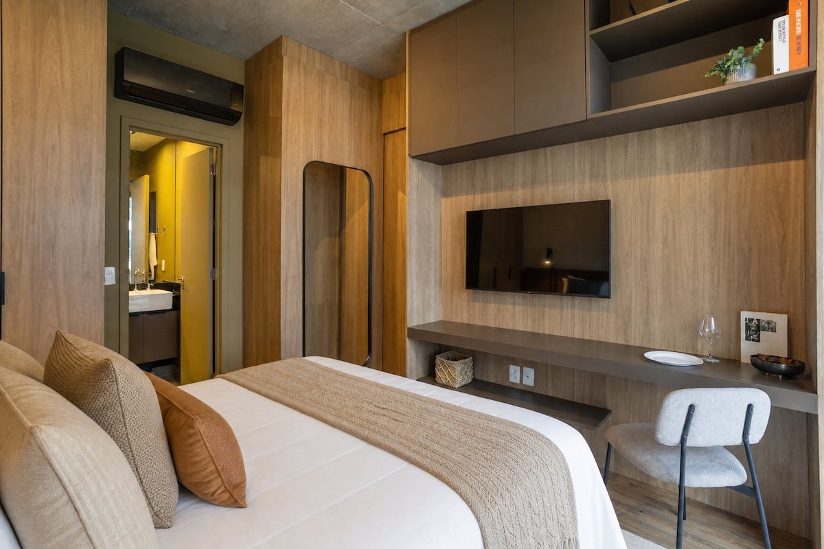 Une chambre d’hôtel moderne, équipée d’un lit, d’un bureau avec une chaise, d’une télévision et d’une salle de bain visible à travers une porte ouverte.