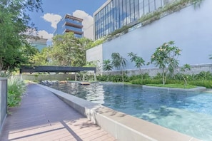 A swimming pool with a wooden deck, surrounded by greenery and modern buildings.