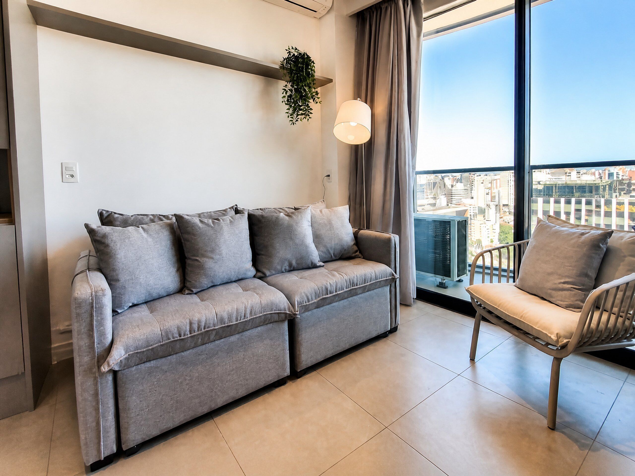 A modern living room with a grey sofa, a single armchair, and a large window offering a city view.