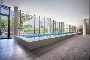 A modern indoor swimming pool with glass walls, wooden flooring, and a view of greenery outside.