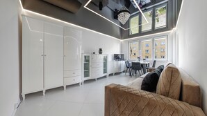 A modern kitchen with white cabinetry, a dining area with a table and chairs, and a large window with a view of buildings. - Apartamenty Sun & Snow 1000-lecia Państwa Polskiego (Wladyslawowo)
