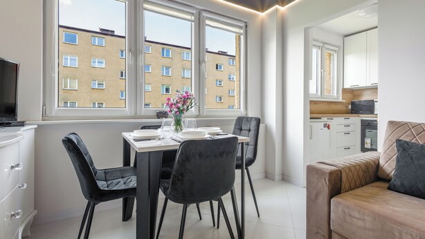 A modern kitchen with white cabinets, a dining area with a table set for two, and a sofa in the living area. - Apartamenty Sun & Snow 1000-lecia Państwa Polskiego (Wladyslawowo)
