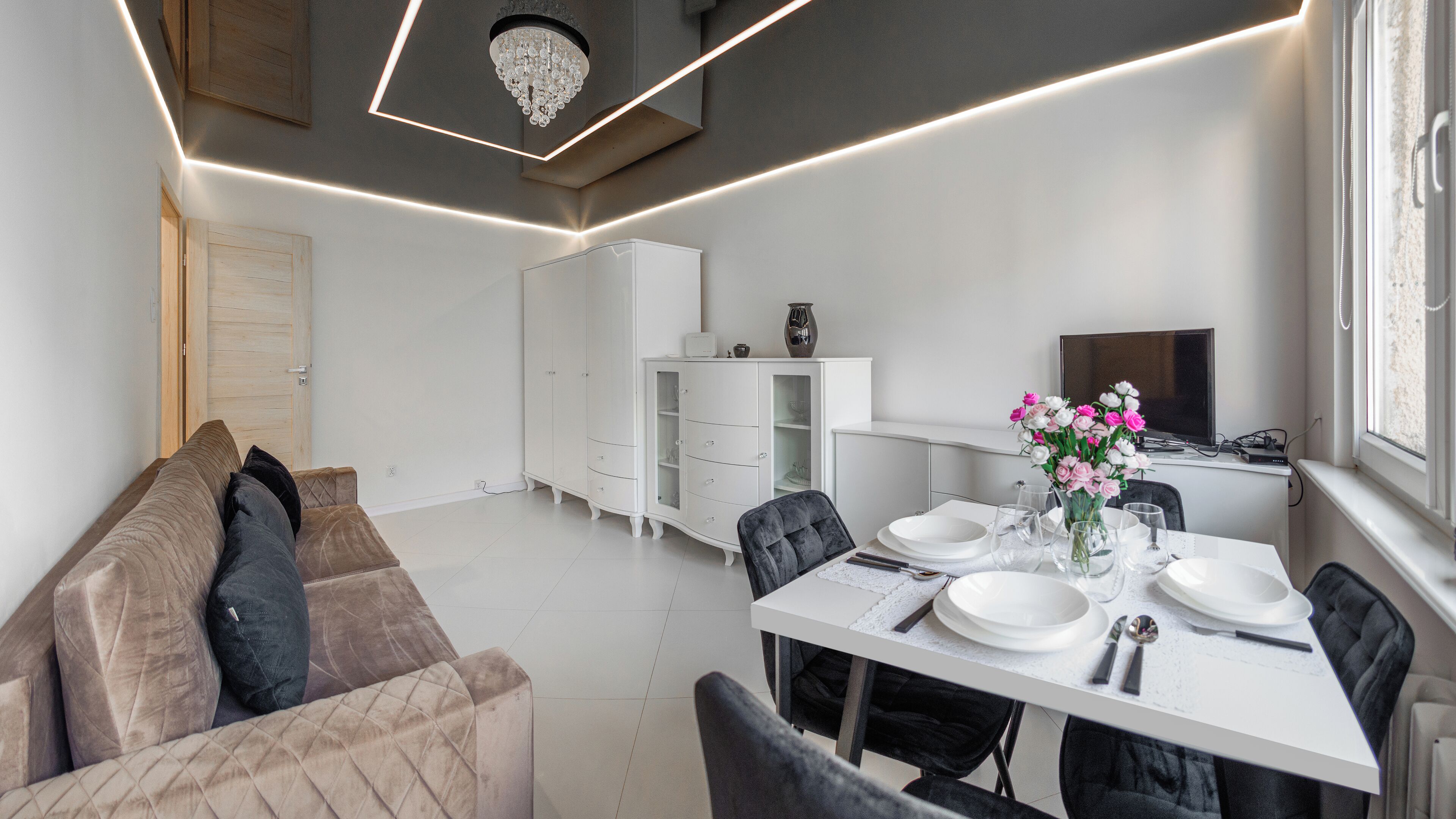 A modern dining area with a white table set for four, a sofa with cushions, a cabinet, and a television on a stand.