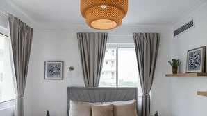 Une chambre à coucher avec un lit, des rideaux, un luminaire suspendu, une fenêtre et une petite plante posée sur une table de chevet.