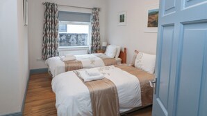 A hotel room with two beds, a window with curtains, a radiator, a bedside table with a lamp, and a door.