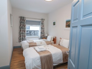 A hotel room with two beds, a window with curtains, a radiator, a bedside table with a lamp, and a door.