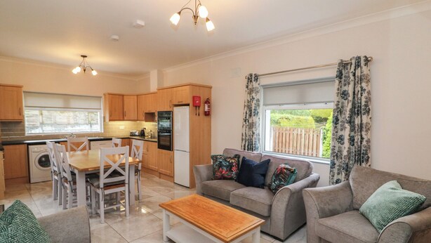 A modern kitchen with wooden cabinets, a dining area with white chairs, a grey sofa set, and a wooden coffee table.