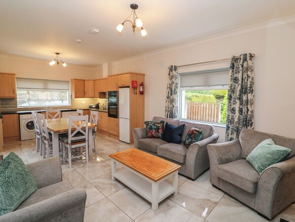 A modern kitchen with wooden cabinets, a dining area with white chairs, a grey sofa set, and a wooden coffee table.