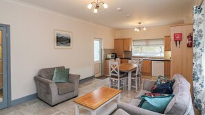 A living room with a sofa, a coffee table, and a dining area with chairs and a table. There is a kitchen with cabinets and a window.