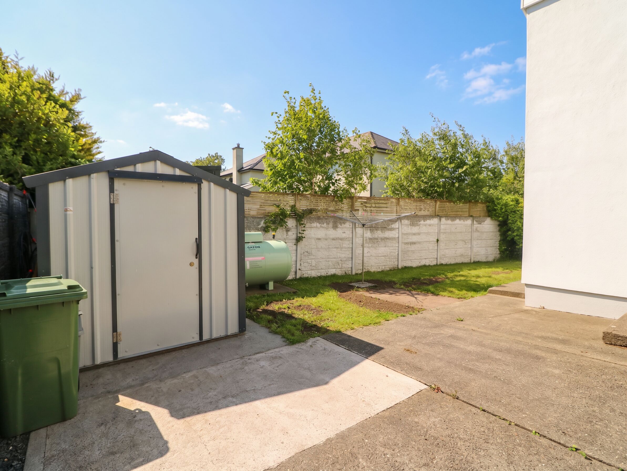 A residential backyard with a shed, a green trash bin, a metal tank, and a concrete path.