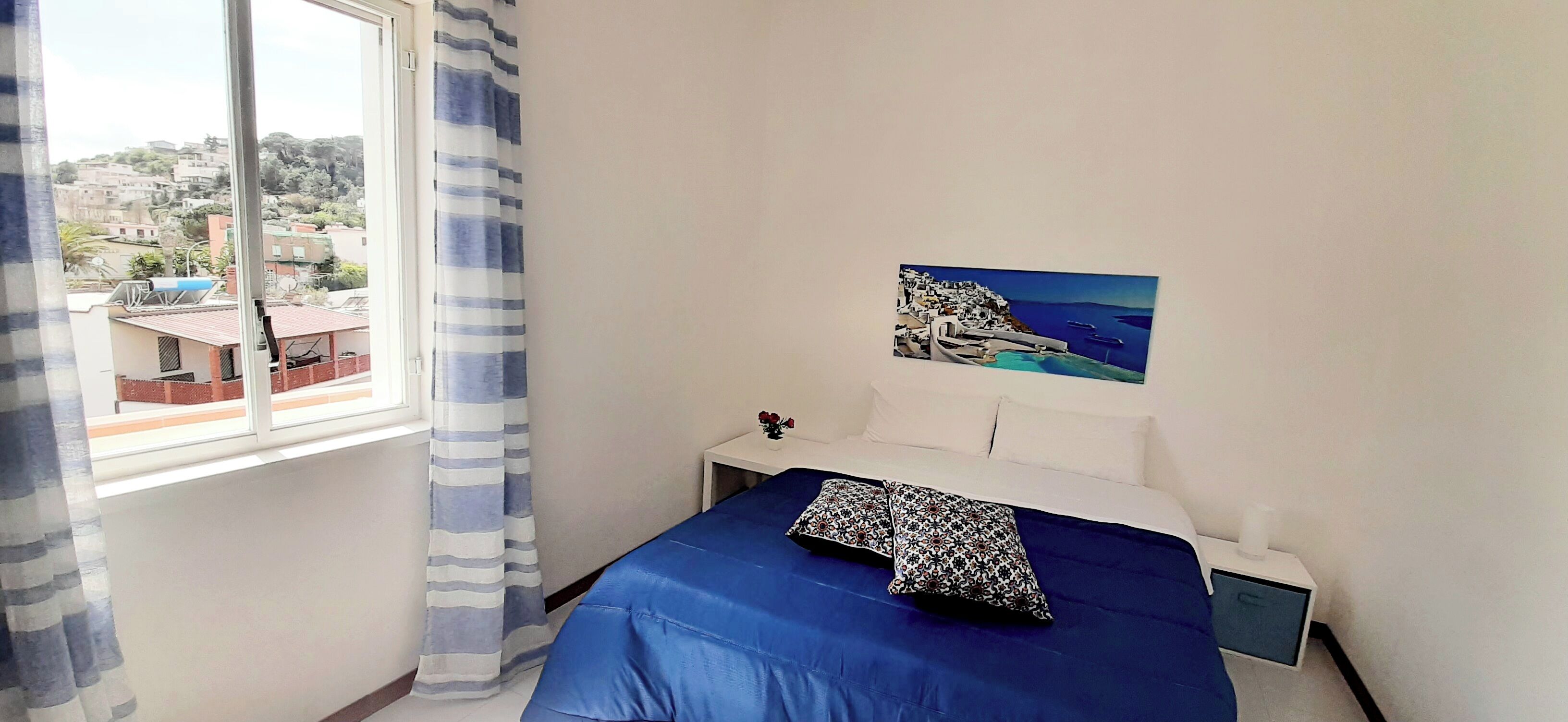 Un dormitorio con cama, mesita de noche, ventana con cortinas y un cuadro de una escena costera.