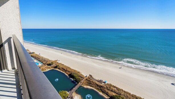 Blick auf einen Strand mit blauem Himmel, feinem Sand und einer kleinen Poolanlage mit Holzterrasse und Stufen zum Wasser.