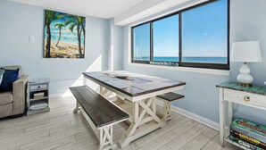 Ein Zimmer mit großem Fenster mit Blick auf den Strand, einem Holztisch, einer Bank, einer Lampe und einem Bücherregal.