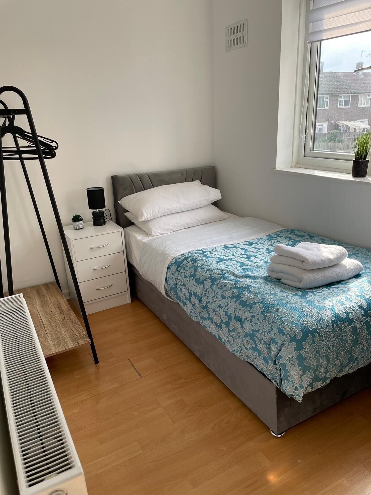 A bedroom with a bed, a bedside table, a radiator, a window with a view, and a plant on the windowsill.