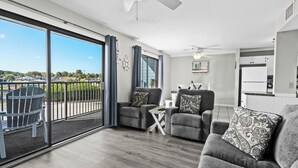 Un vaste espace de vie avec balcon, des canapés gris ornés de coussins à motifs, une petite table blanche et un ventilateur de plafond.