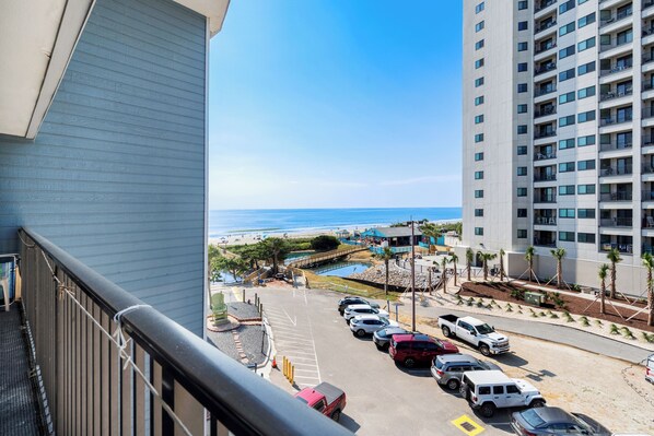 A view from a balcony overlooking a parking area with several cars, a beach, and a building with multiple balconies.