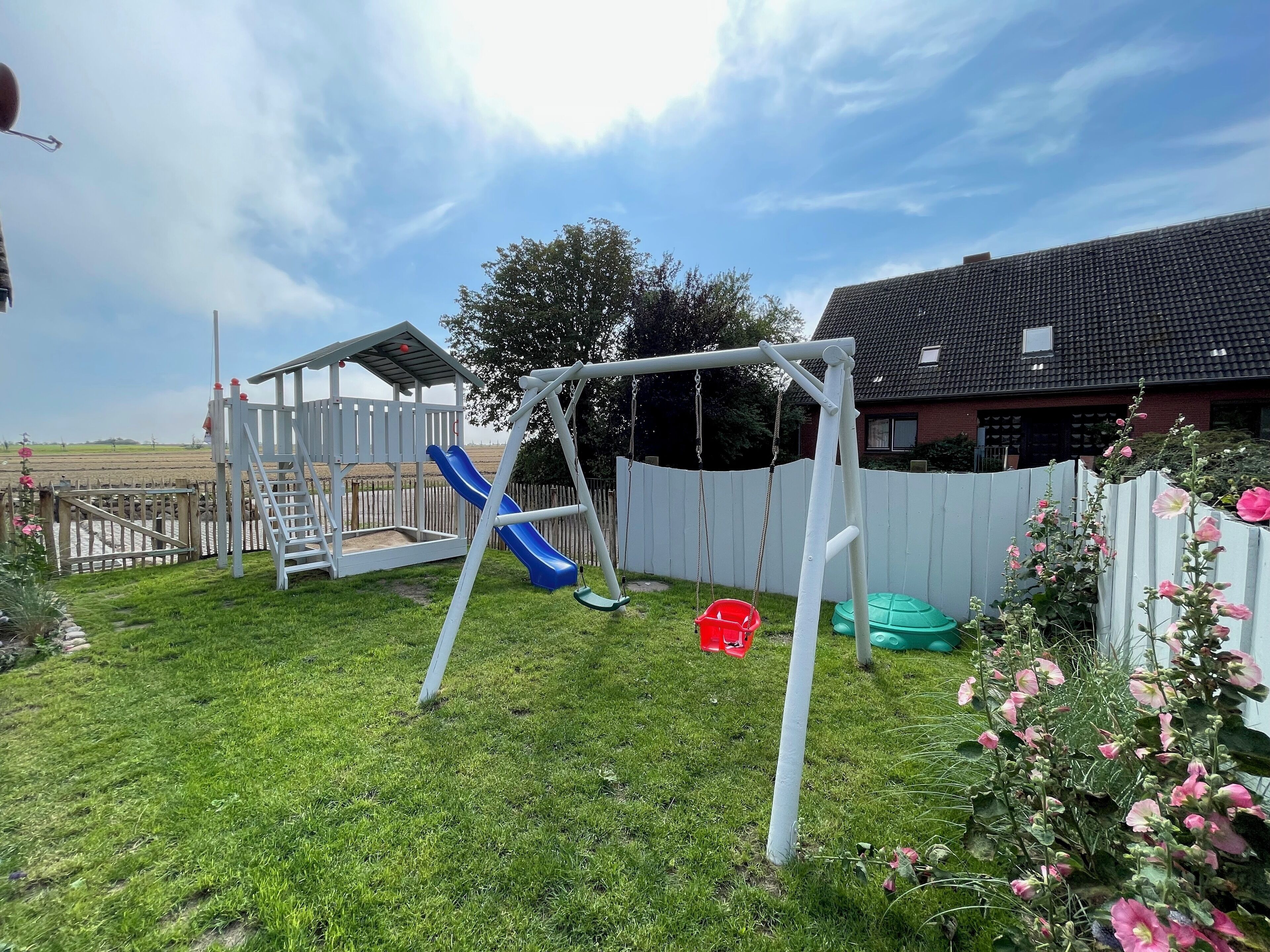 A backyard with a swing set, a slide, a wooden playhouse, and a fenced area.