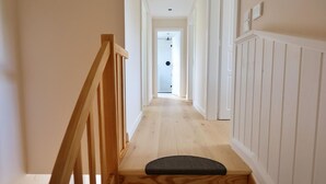 A wooden staircase with a black mat at the bottom, leading to a hallway with white walls and wooden flooring. - Island idyll Villa Kunterbunt (Fehmarn)