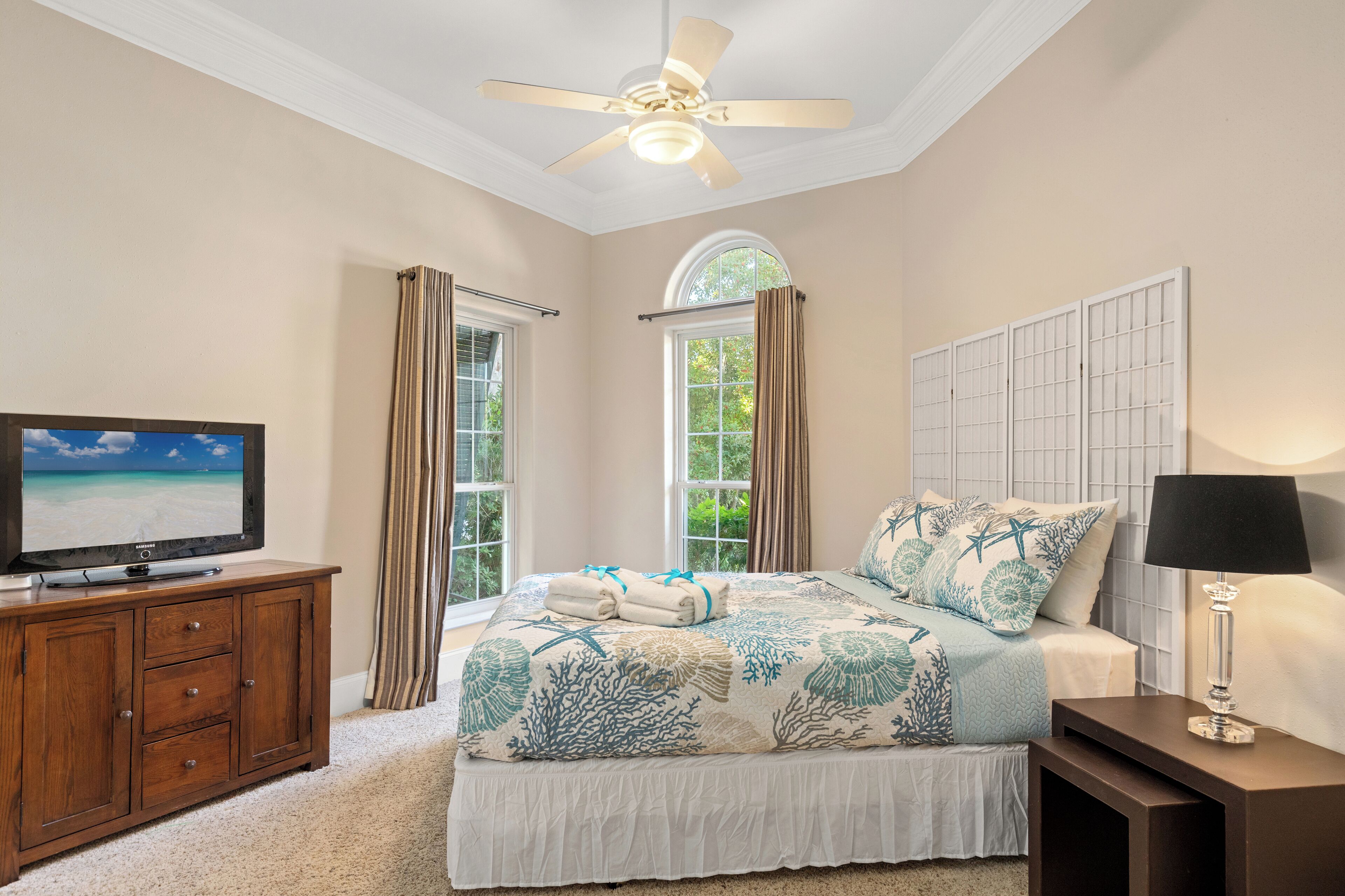 A bedroom with a bed, a TV on a wooden cabinet, a ceiling fan, and a window with curtains.