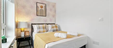 A neatly made bed with a white and yellow bedspread, a bedside lamp, and a framed picture on the wall.