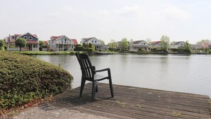 Vista del lago con una silla en una terraza de madera, casas al fondo y un seto en primer plano.