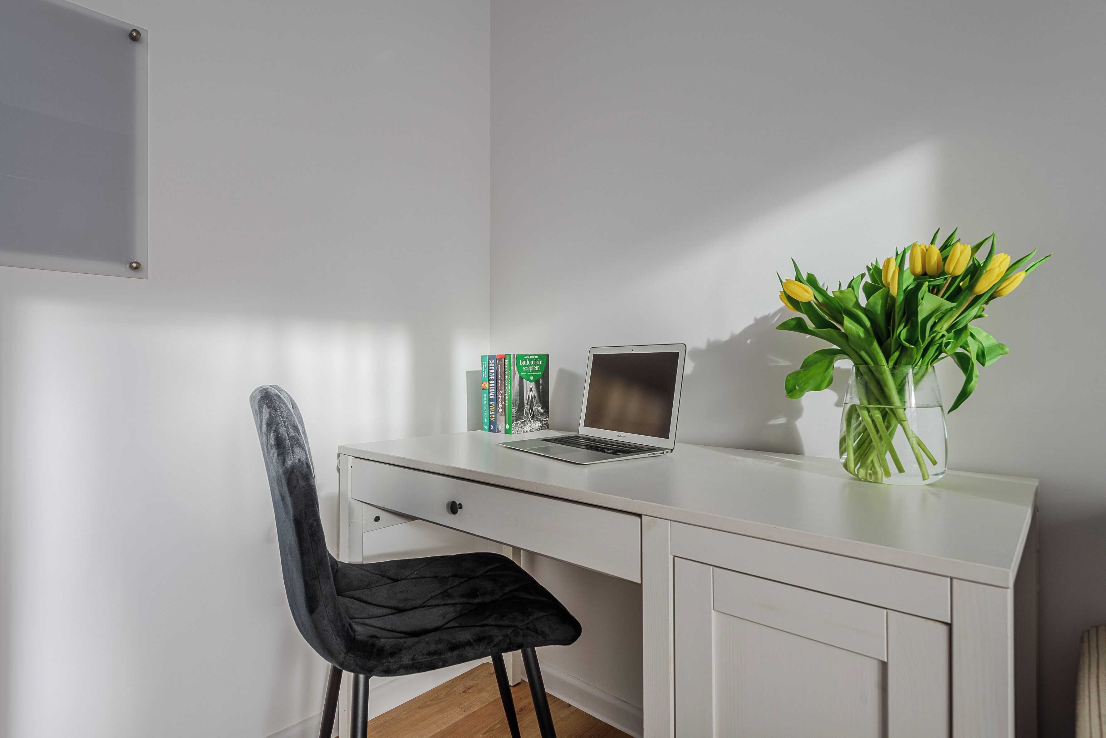 Un escritorio blanco con una computadora portátil, una silla negra y un florero con flores amarillas.