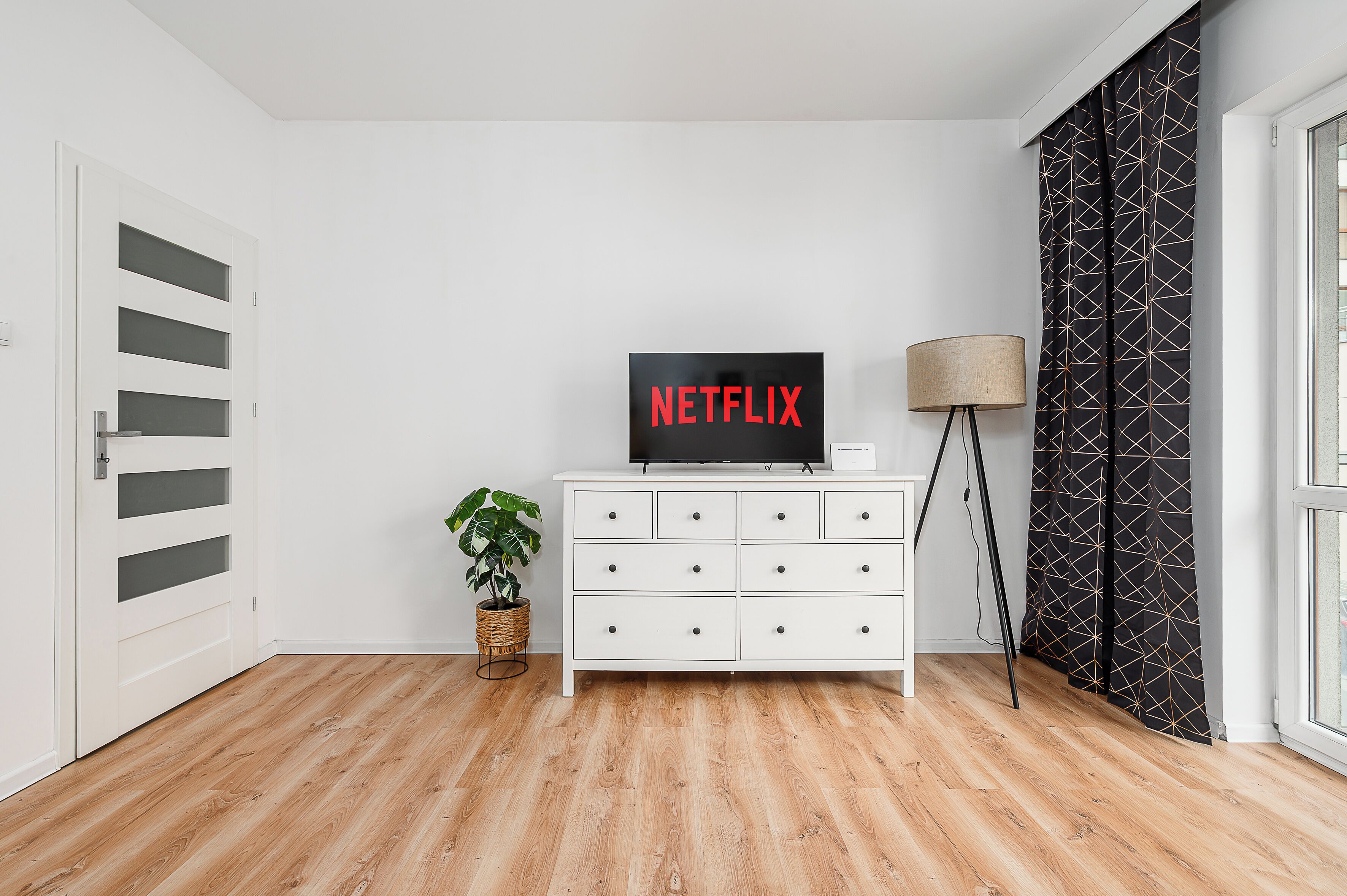 Huoneessa on valkoinen lipasto, viherkasvi, Netflixin logo ja lamppu.