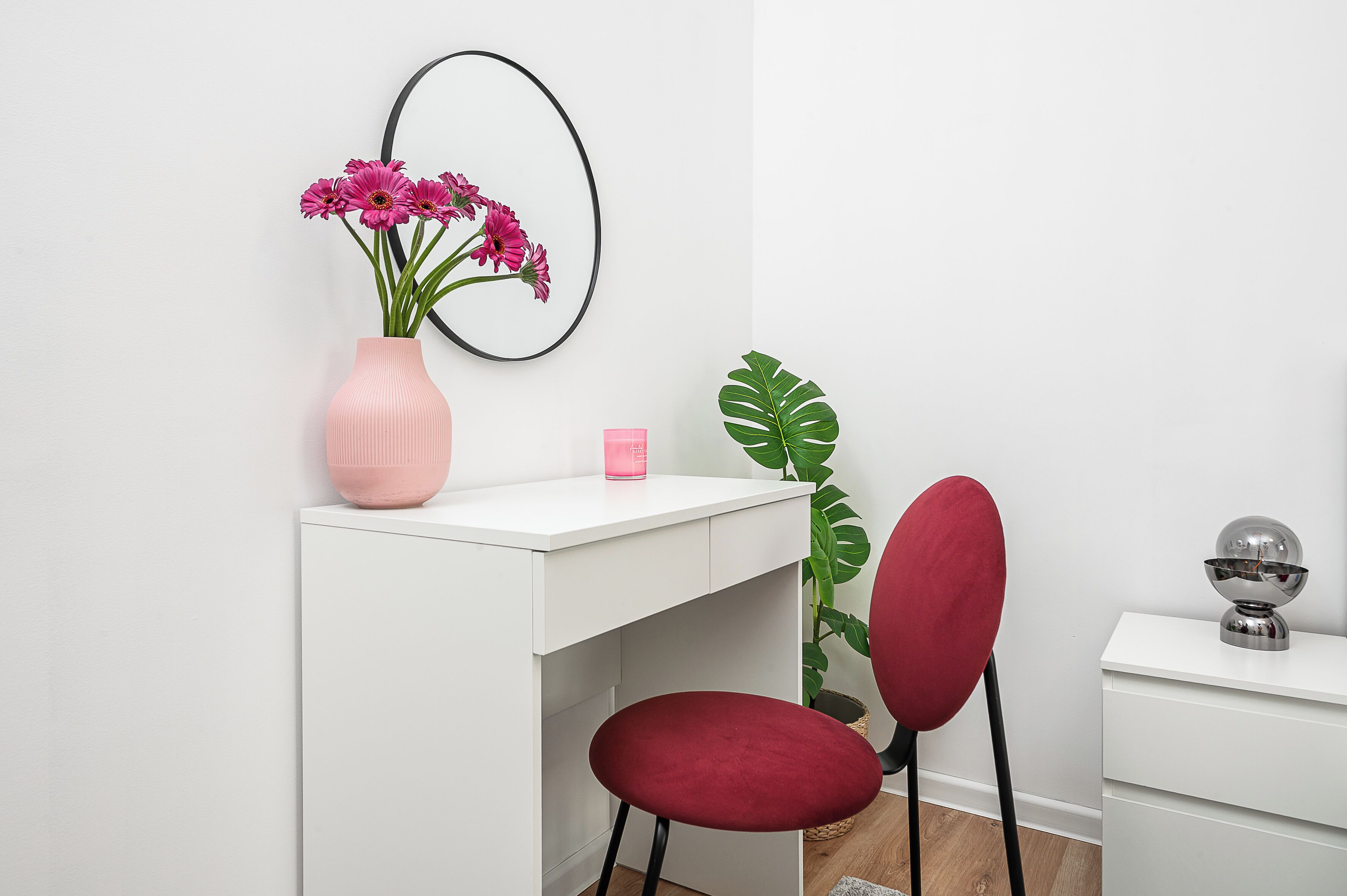 A white desk with a pink vase of flowers, a red chair, a large green plant, and a small white nightstand with a globe.
