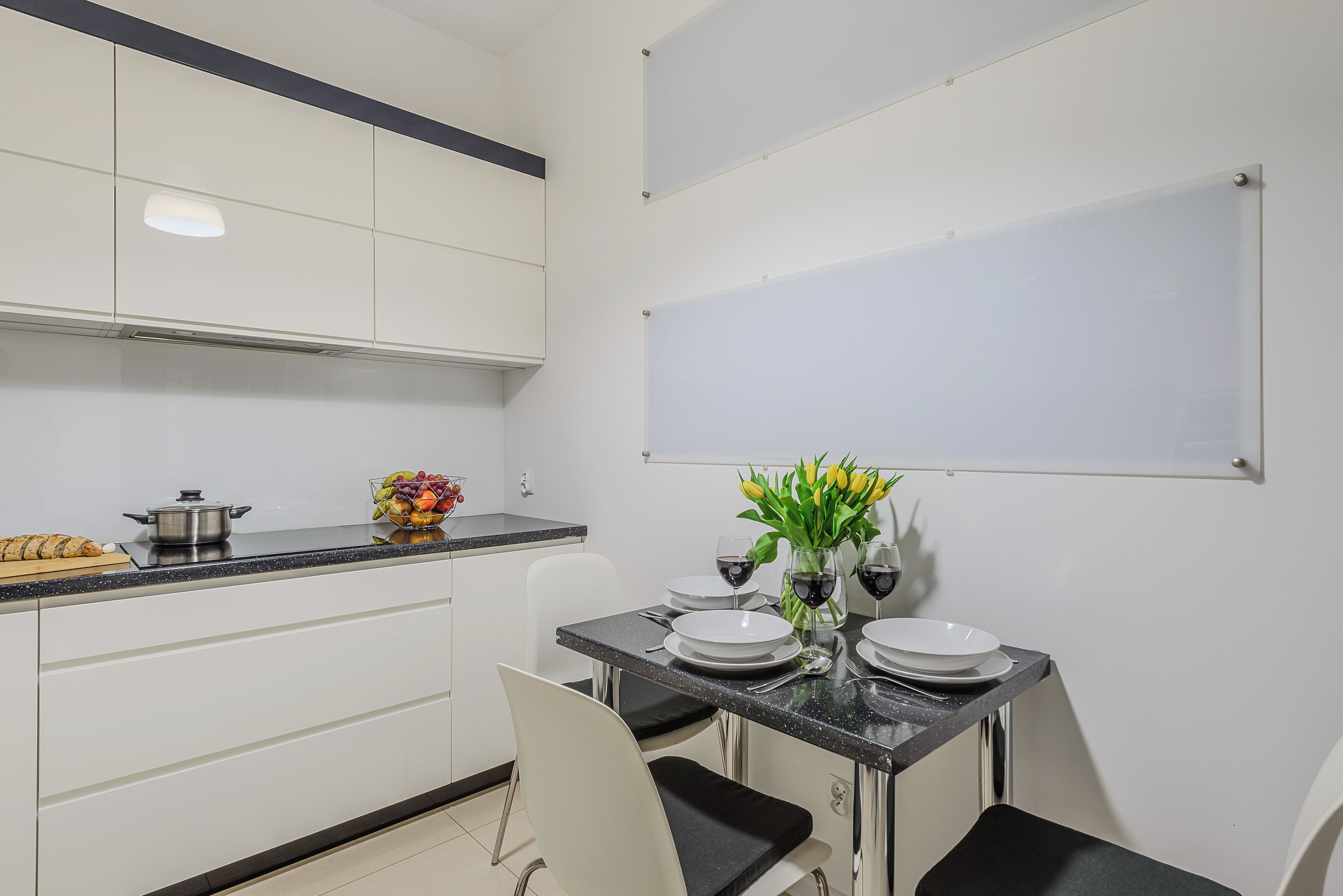 A modern kitchen with white cabinets, a black countertop, and a dining table set with plates, wine glasses, and a vase of flowers.