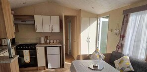 A compact kitchen with a stove, oven, and dishwasher, adjacent to a dining area with a table and chairs. - Seaside Serenity Cabin Retreat (Rhyl)
