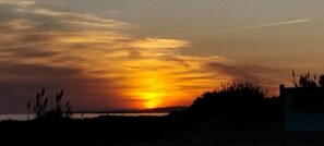 Un atardecer sobre un cuerpo de agua, con siluetas de vegetación y un edificio al frente.